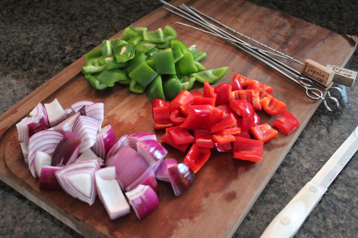 Diced Onions and Bell Peppers on a wooden cutting board next to metal skewers.