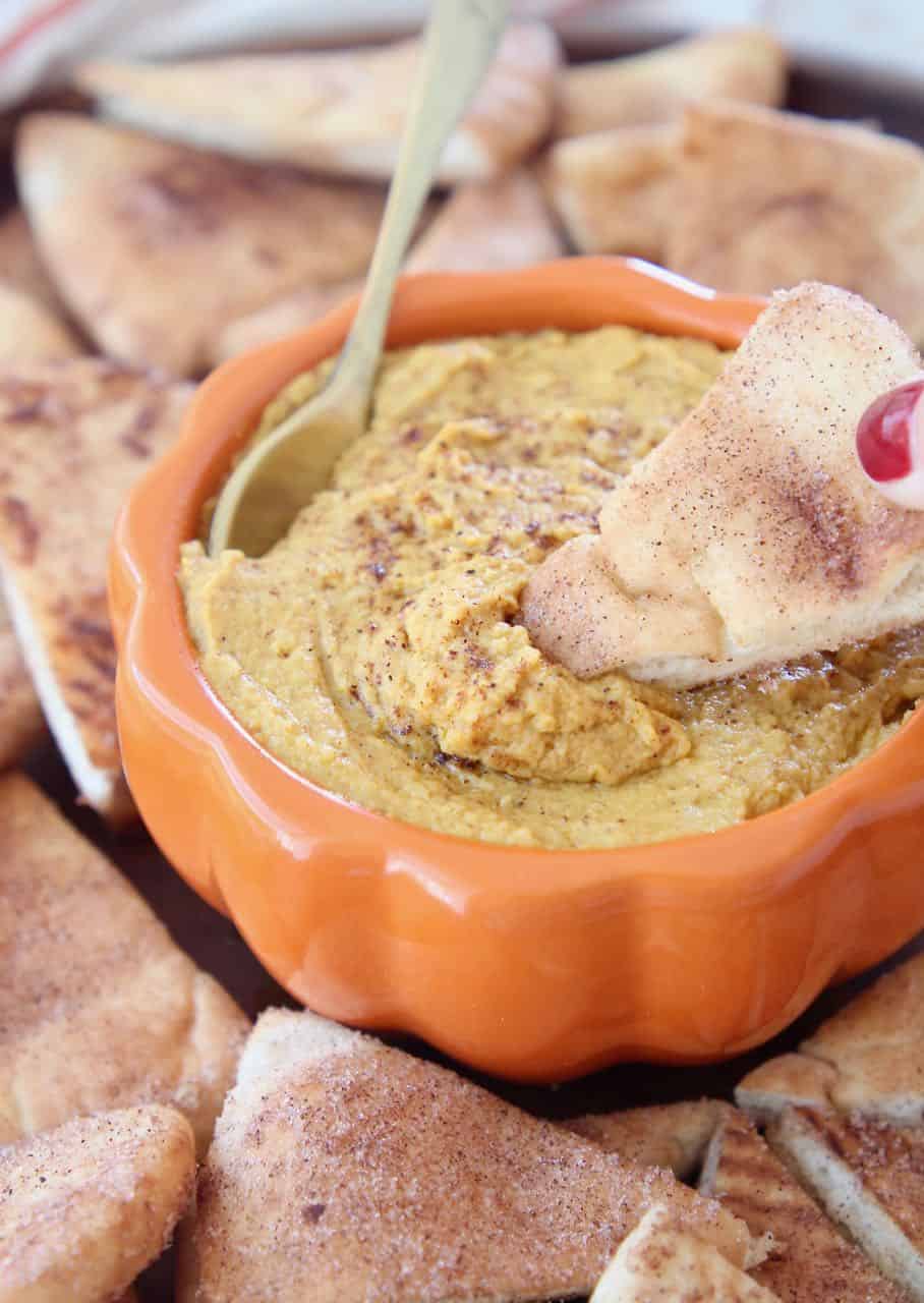 Pumpkin hummus in orange bowl with pita chip being dipped in the hummus