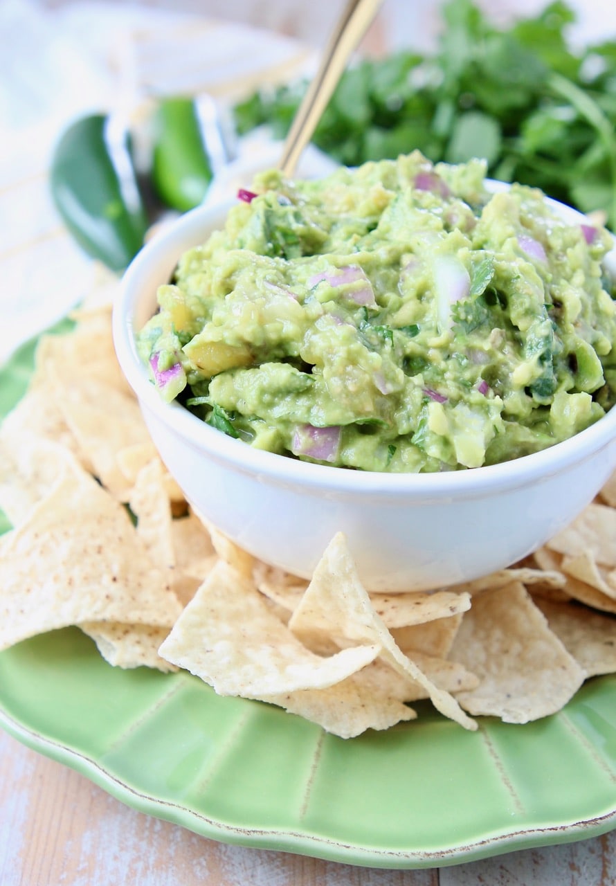 Tomatillo guacamole in white bowl on green plate, surrounded by tortilla chips