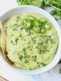Creamy green salsa in bowl with gold spoon and serrano peppers