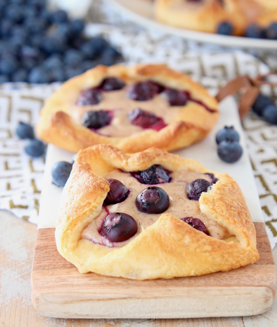 Two blueberry pastries sitting on a wood and marble cutting board with fresh blueberries around and behind them
