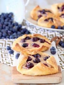 Blueberry Pastries sitting on cutting board on top of gold and white patterned towel with a white colander of blueberries spilling on in the background next to a plate of blueberry pastries