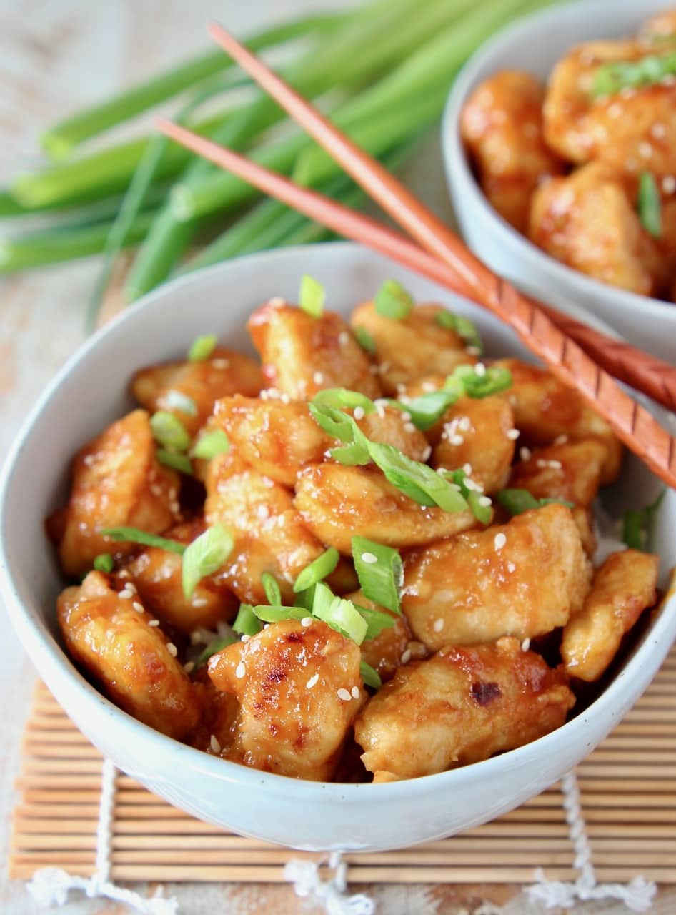 Orange chicken in bowl topped with sliced scallions and sesame seeds, with wood chopsticks on the side of the bowl