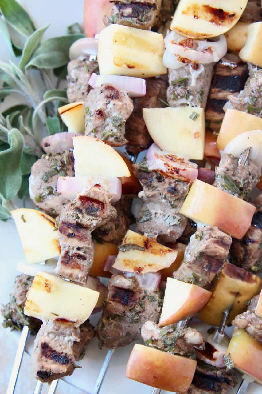 Pork tenderloin skewers with apples and shallots, stacked up on a plate with fresh herbs