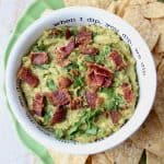 Guacamole in white bowl topped with cooked bacon, with tortilla chips on the side