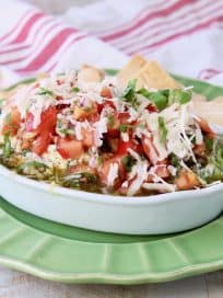 Italian 5 layer dip topped with tomato basil bruschetta in a white serving bowl sitting on a green plate with pita chips