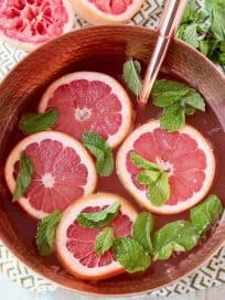 Spiced rum punch in copper bowl, topped with fresh grapefruit slices and fresh mint leaves, with rose gold ladle and fresh mint leaves on the side