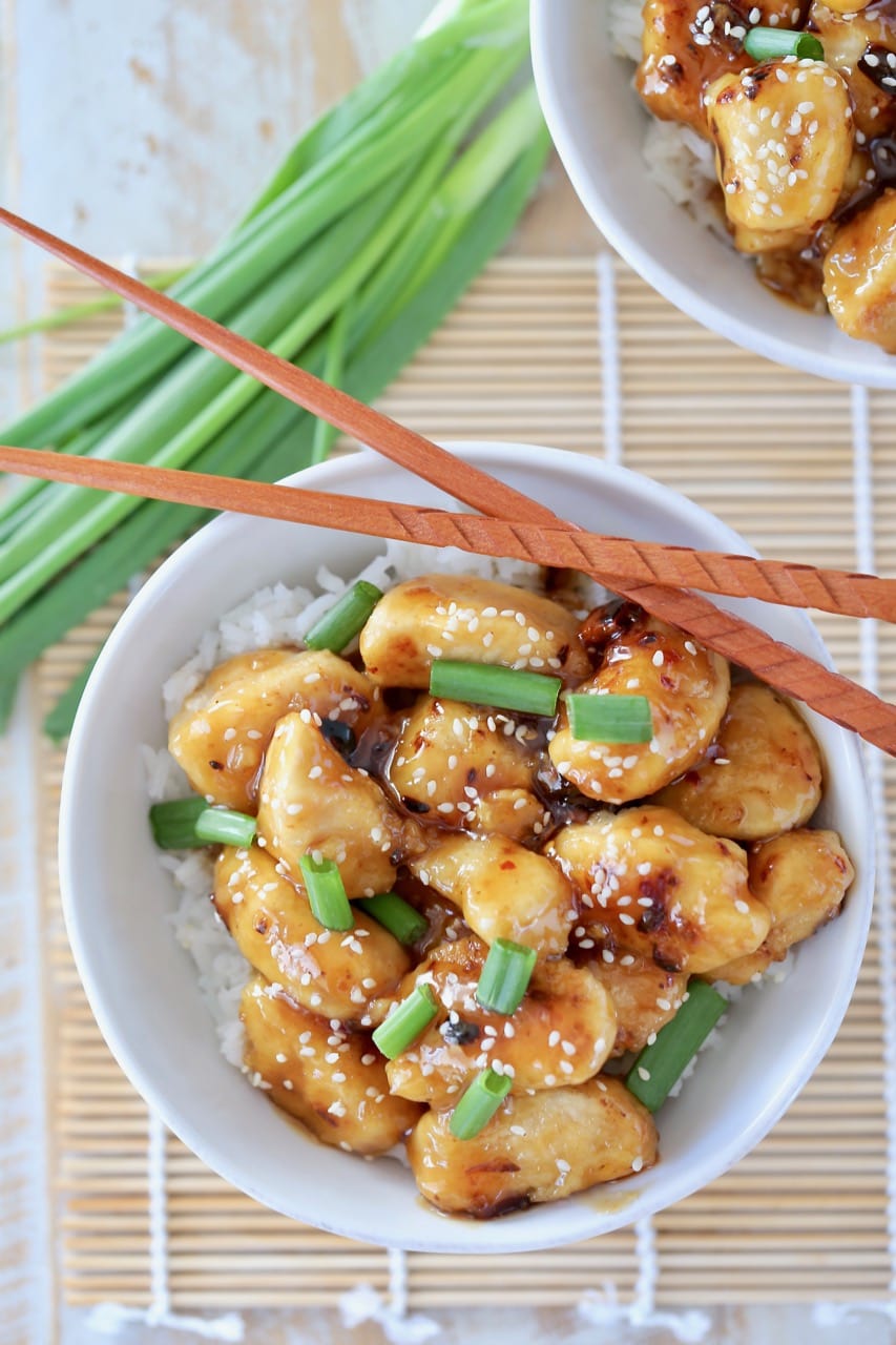 Overhead shot of Chinese honey chicken in white bowl with white rice, green onions and sesame seeds with chopsticks on the side of the bowl and green onions next to the bowl