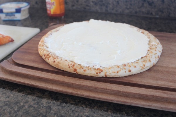 A cooked pizza crust covered with a cheese sauce on a wooden cutting board. 