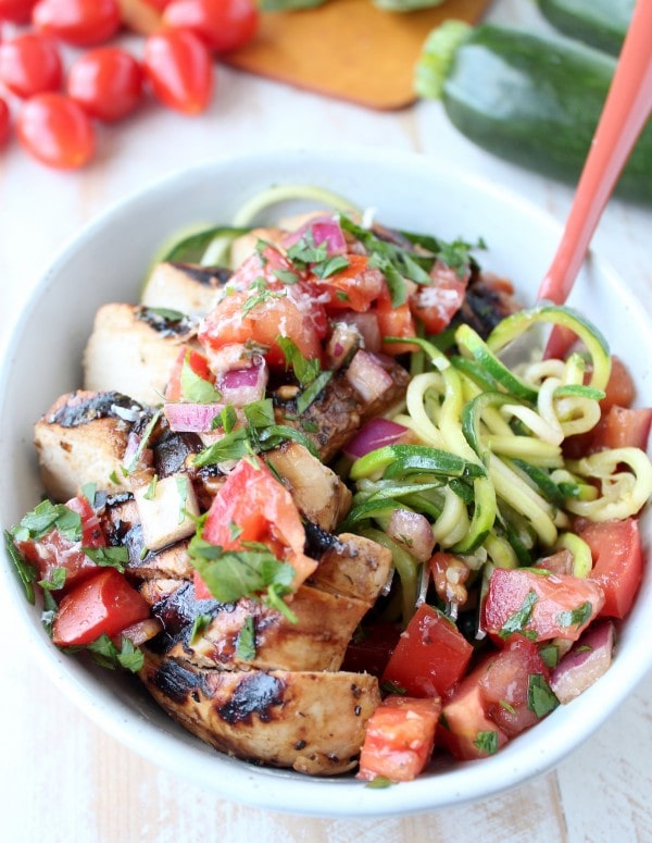 Zucchini noodles spun around a fork in a bowl with tomato basil bruschetta and grilled chicken