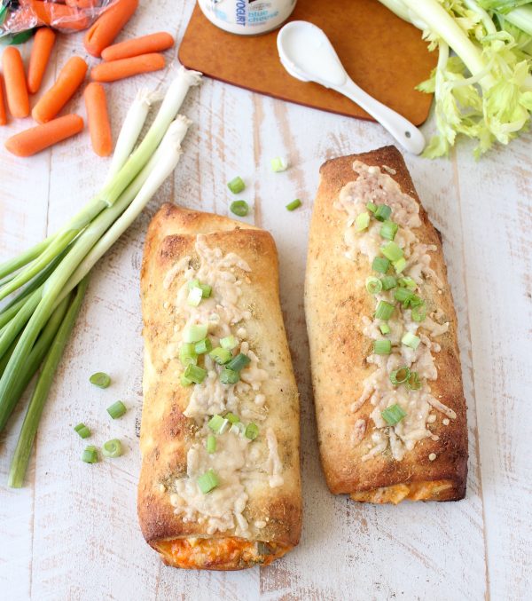 Two cooked Chicken Bakes on a wooden surface next to carrots and green onions. 