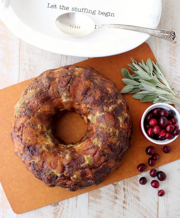 Stuffing that has been cooked in a bundt pan on a wooden surface surrounded by a spoon on a white plate, herbs, and cranberries.