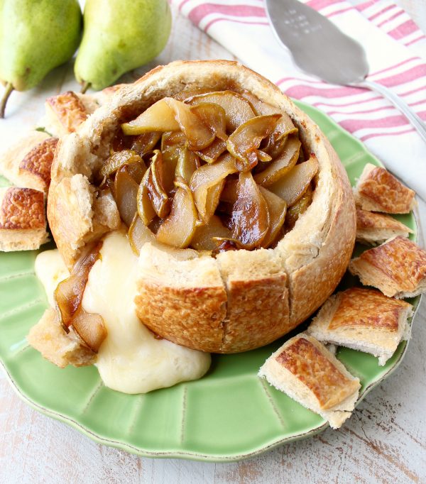 Baked brie spilling out of bread bowl onto green plate with caramelized pears on top