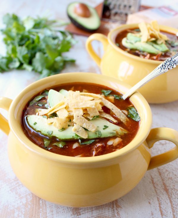 A yellow bowl filled with enchilada soup topped with slices of avocado and tortilla strips with a second bowl in the background.