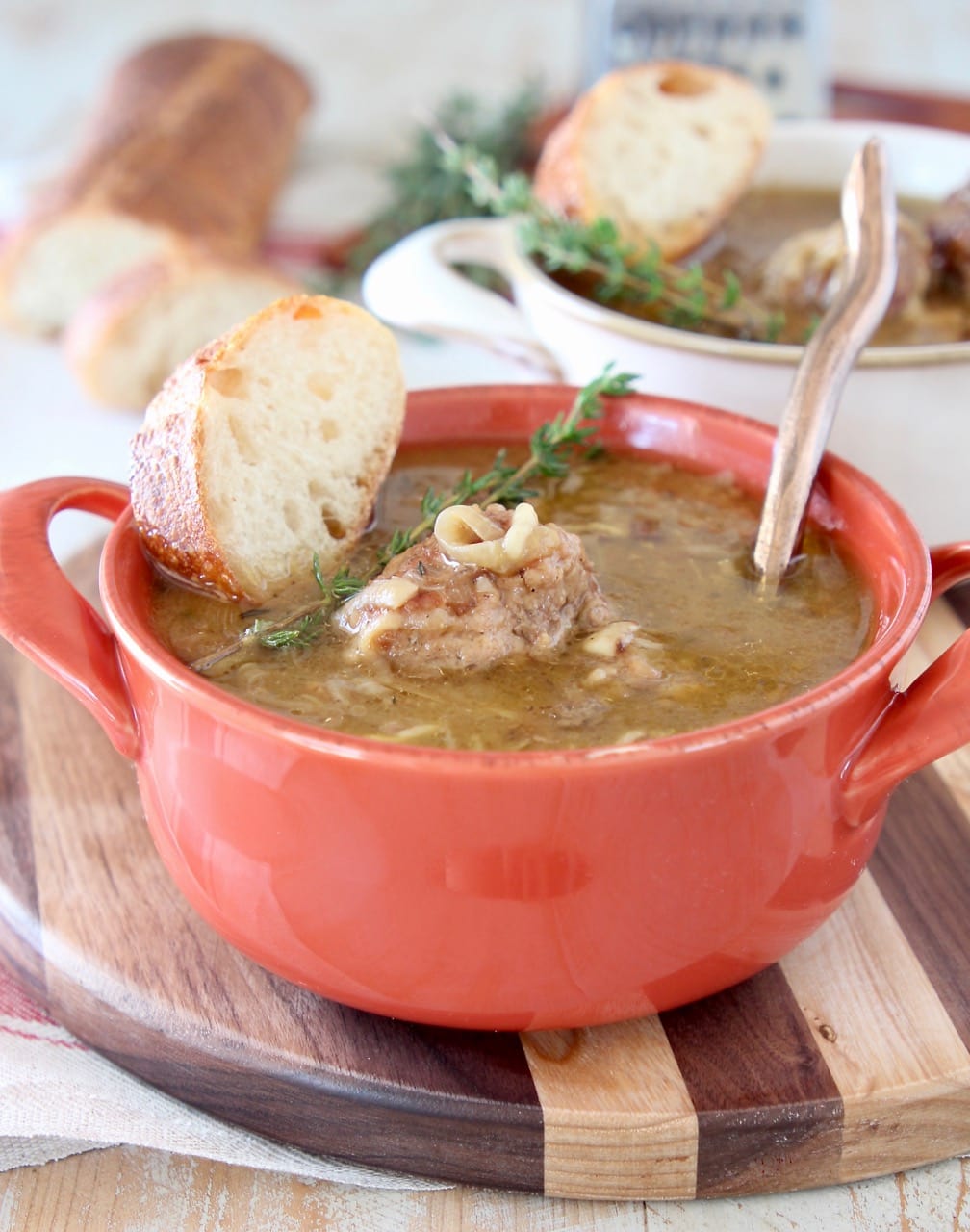 French onion meatball soup in orange crock with fresh spring of thyme on top and copper spoon in the soup with a slice of bread on the side, sitting on a wood striped cutting board