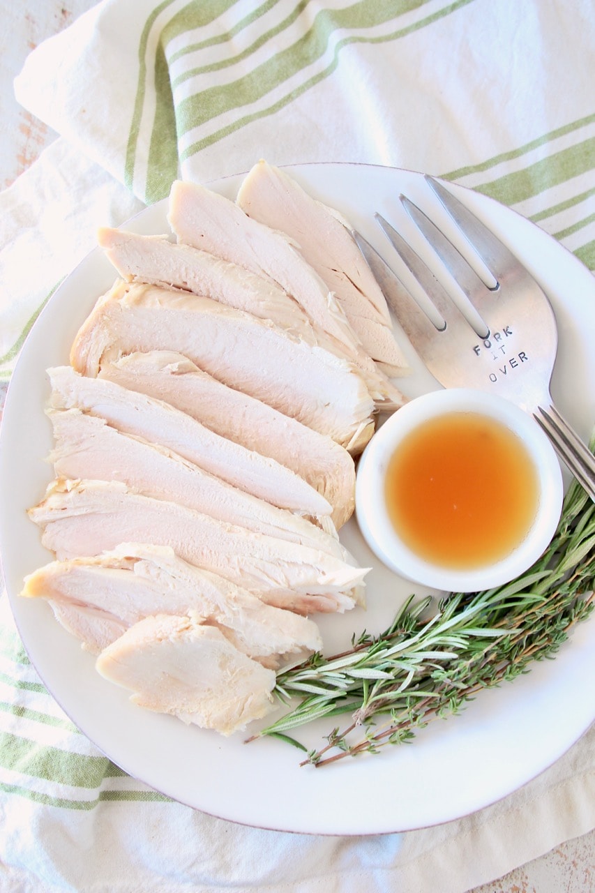 Sliced turkey breast on plate with small white bowl of apple reduction, large fork and fresh rosemary and thyme sprigs, on white and green cloth towel