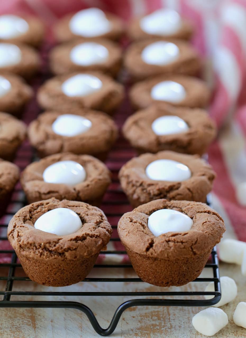 Hot chocolate cookies made into cups and filled with marshmallow creme on black wire rack with mini marshmallows on the side sitting on red and white striped towel
