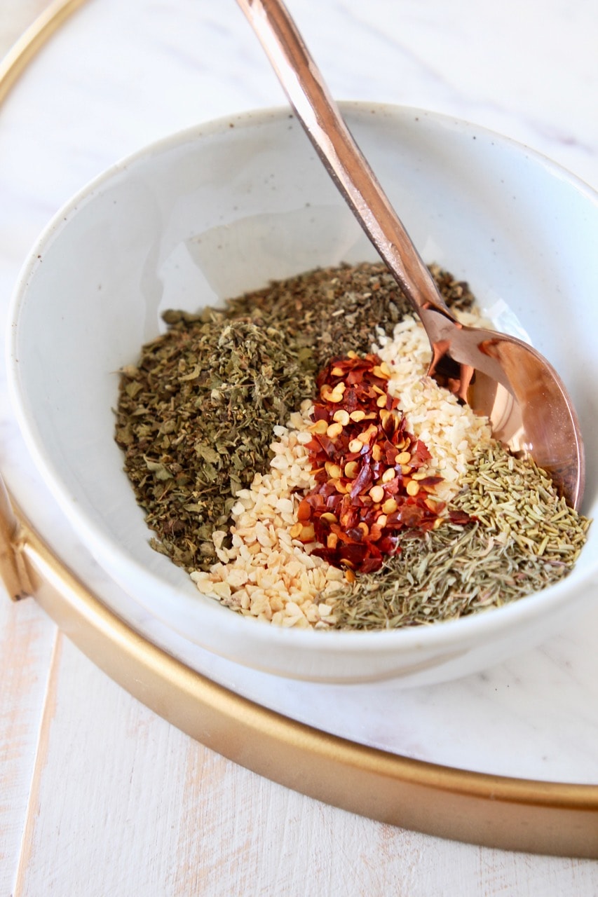 Italian seasoning blend of different herbs and spices together in a white bowl with a copper spoon, sitting on a white marble cutting board