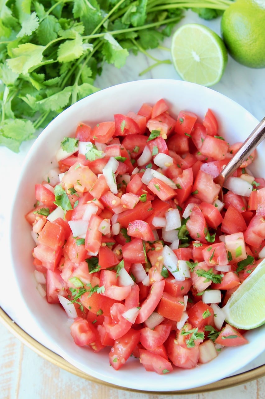 Overhead shot of pico de gallo in white bowl with fresh lime wedges and spoon in the bowl, with fresh cilantro on the side