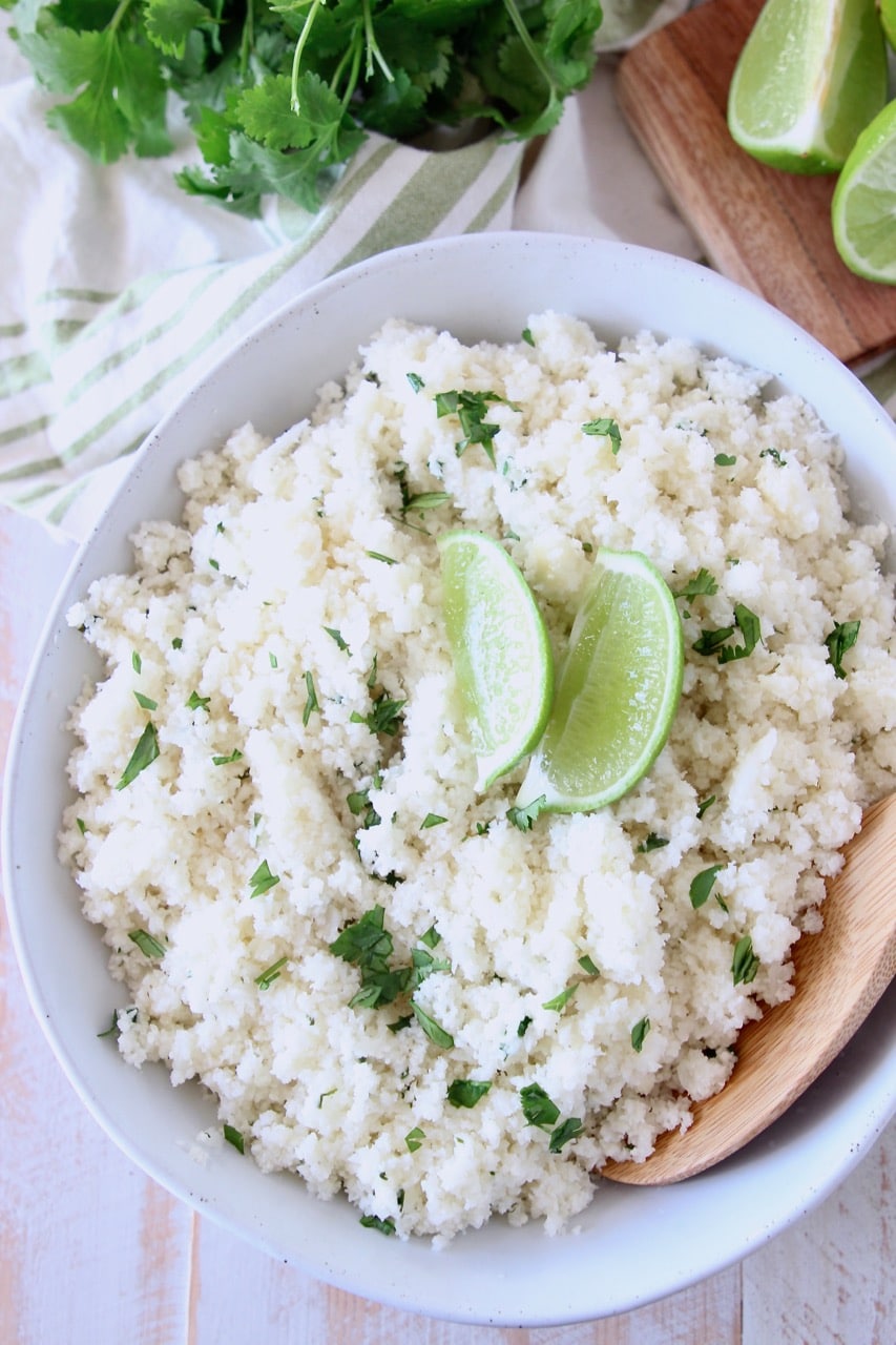 Large white bowl of cilantro lime cauliflower rice with two lime wedges on top and wooden spoon in the bowl