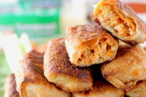 Buffalo chicken egg rolls piled up on a green plate with one egg roll cut in half on top, with cheddar cheese and a cheese grater in the background with text overlay "buffalo chicken egg rolls, quick & easy appetizer recipe"