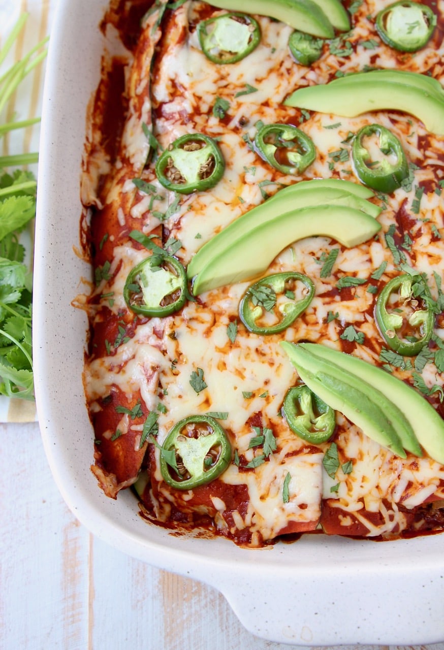 Overhead shot of enchiladas covered in red sauce and cheese in baking dish topped with avocado and jalapenos