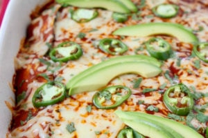 Enchiladas in casserole dish topped with red sauce, jalapenos and avocado