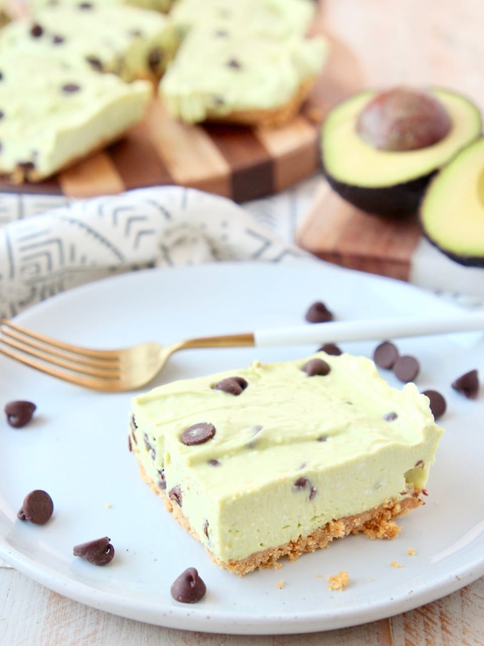 Avocado cheesecake bar with graham cracker crust on white plate with gold fork and chocolate chips