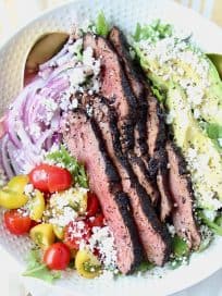 Grilled steak on top of salad in a white bowl with gold salad serving spoons