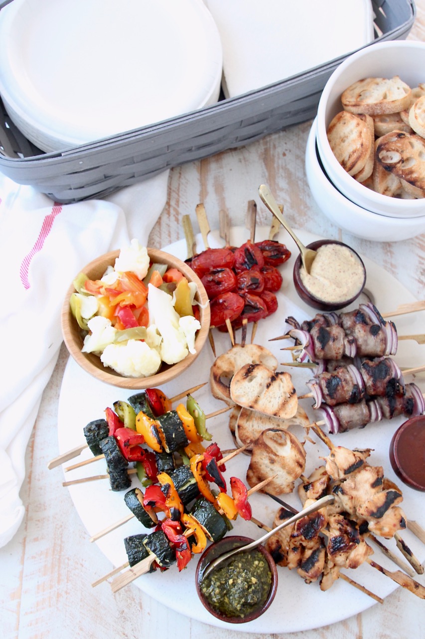 Grilled mini skewers on charcuterie board next to bowl of toasted baguette slices and basket of napkins