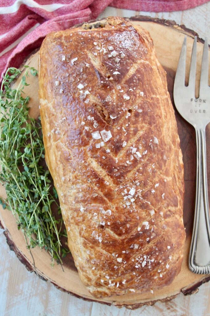 baked beef wellington on wood serving board with fresh herbs and serving fork