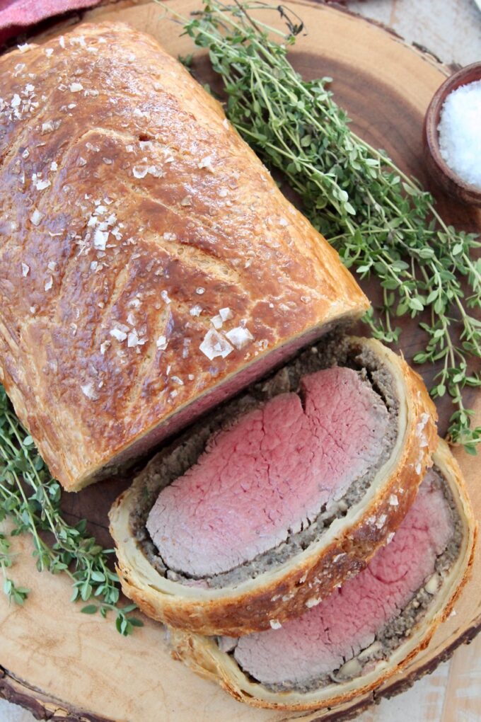 cooked, sliced beef wellington on wood serving tray with fresh herbs