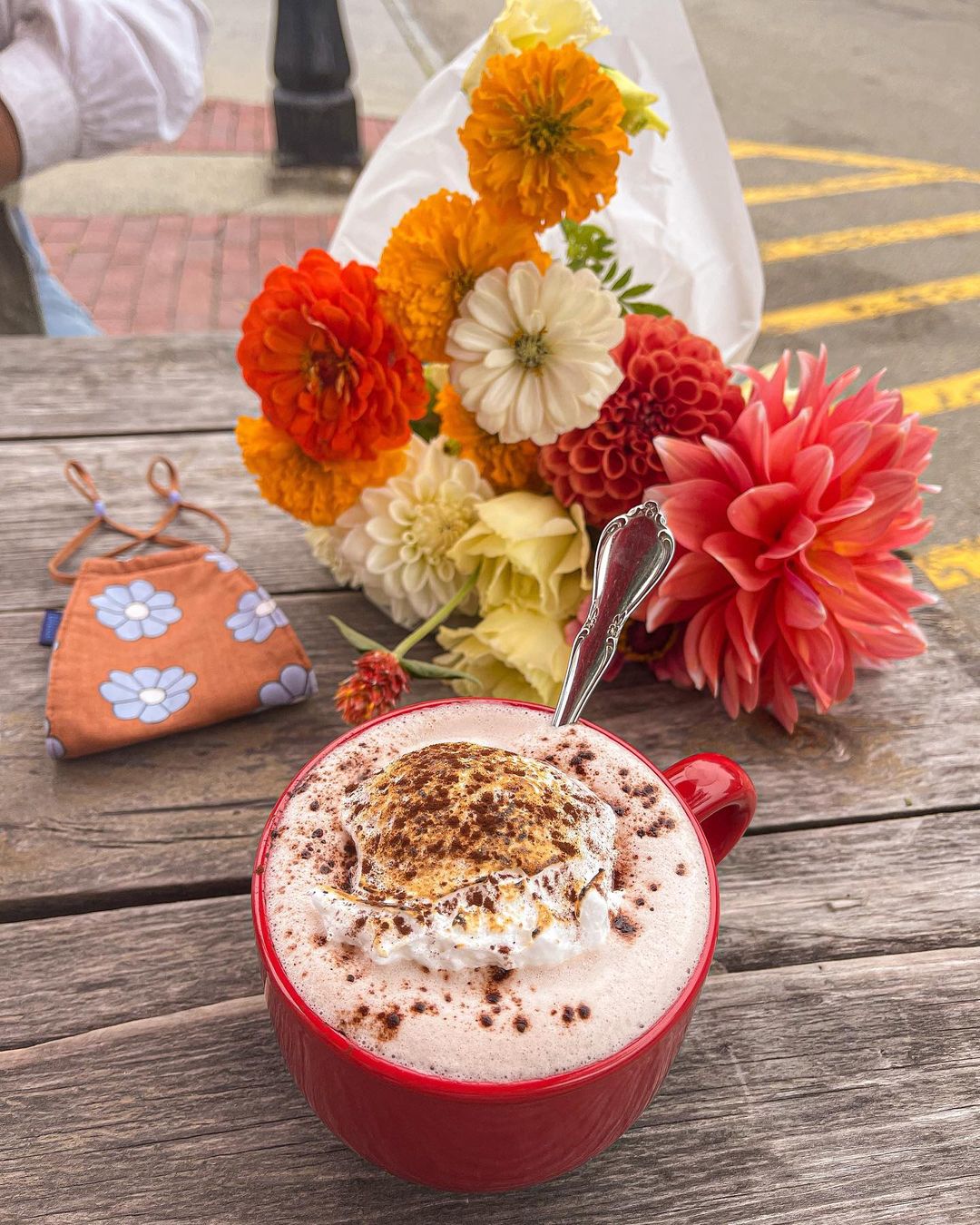 Coffee in a red cup, a bouquet of flowers, and an orange floral face mask on a wooden table