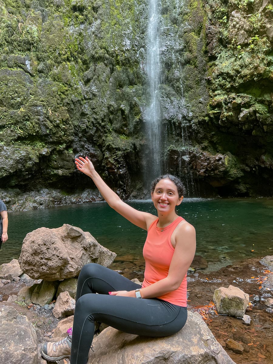 A woman sitting on a rock with the waterfall in the background on a Levada Caldeirao Verde hike