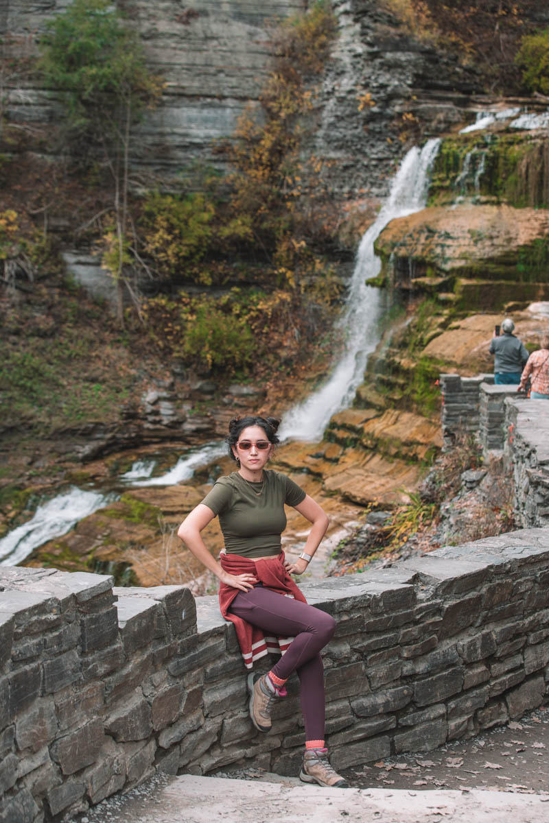 A woman sitting on a rock fence with Lucifer Falls in the background at Robert Treman State Park