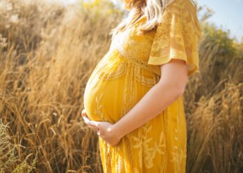 Mujer embarazada vistiendo un vestido floral amarillo de pie mientras toca su barriga y mirando hacia su lado derecho cerca del campo marrón durante el día