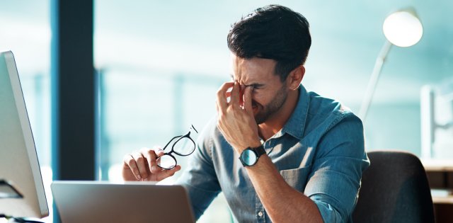 office worker at desk with strained eyes taking off his glasses and massaging the bridge of his nose.