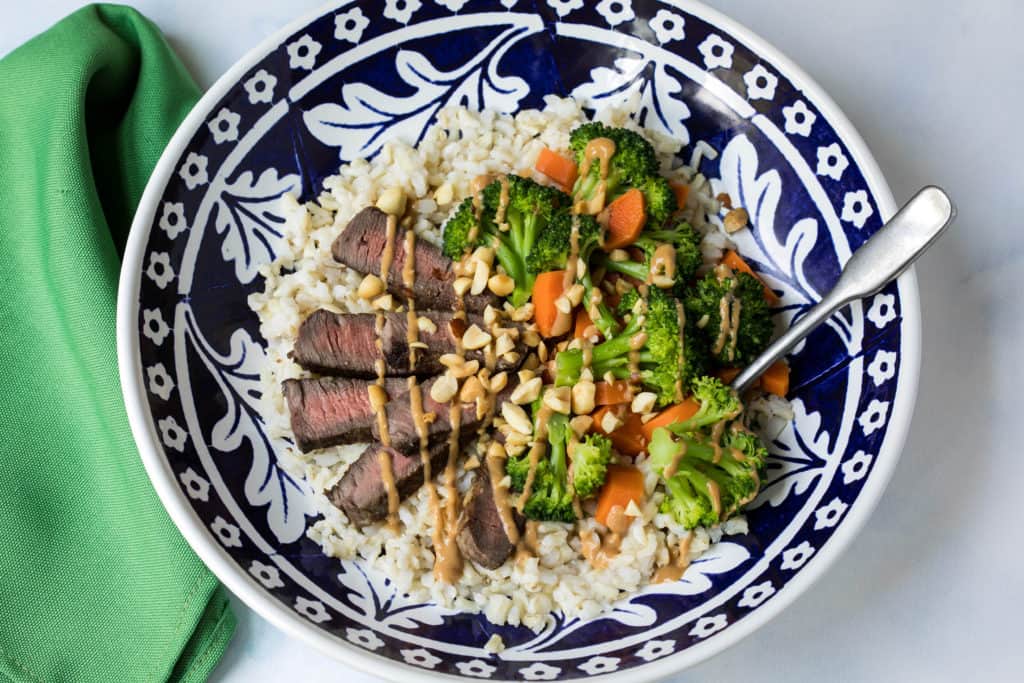 grain bowl with steak, carrots, broccoli and peanut sauce on blue and white plate with green napkin