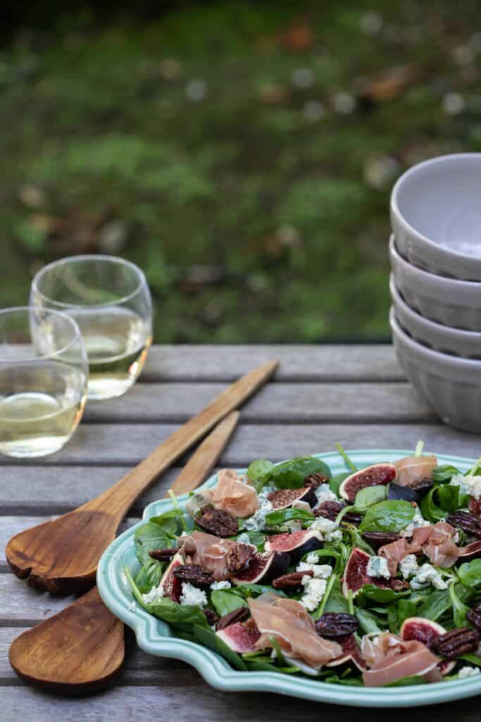 fig salad with blue cheese, pecans, and proscuitto on wooden table outside with glasses of wine, bowls, and serving utensils