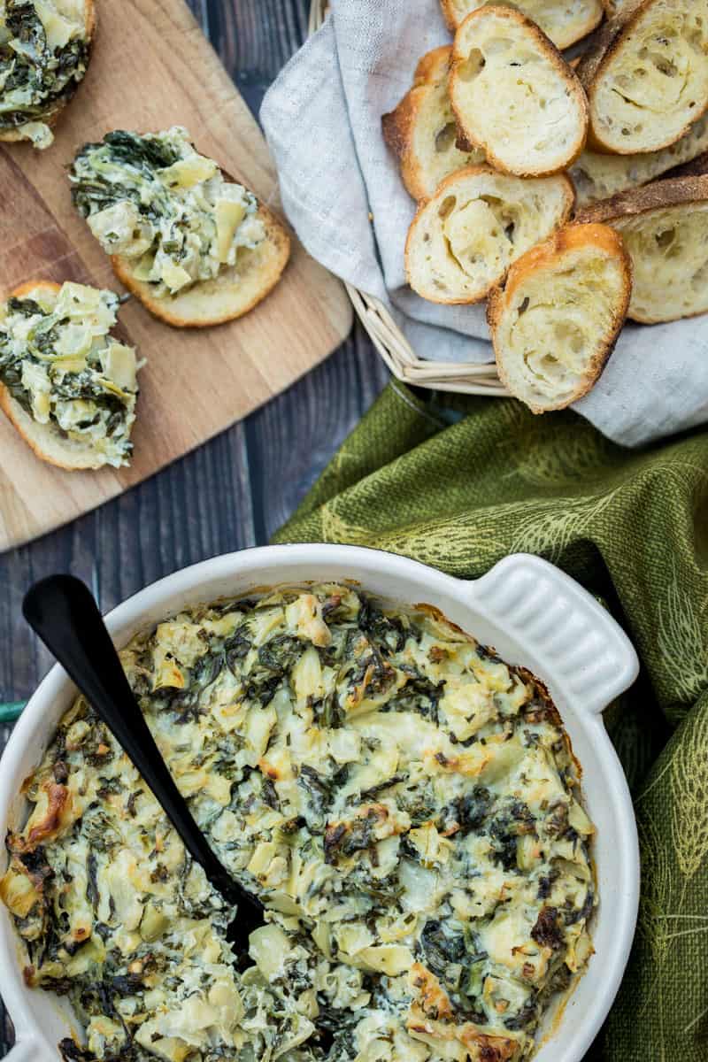 dish of healthier baked spinach artichoke dip  with napkin, basket of crostini and crostini with dip on cutting board