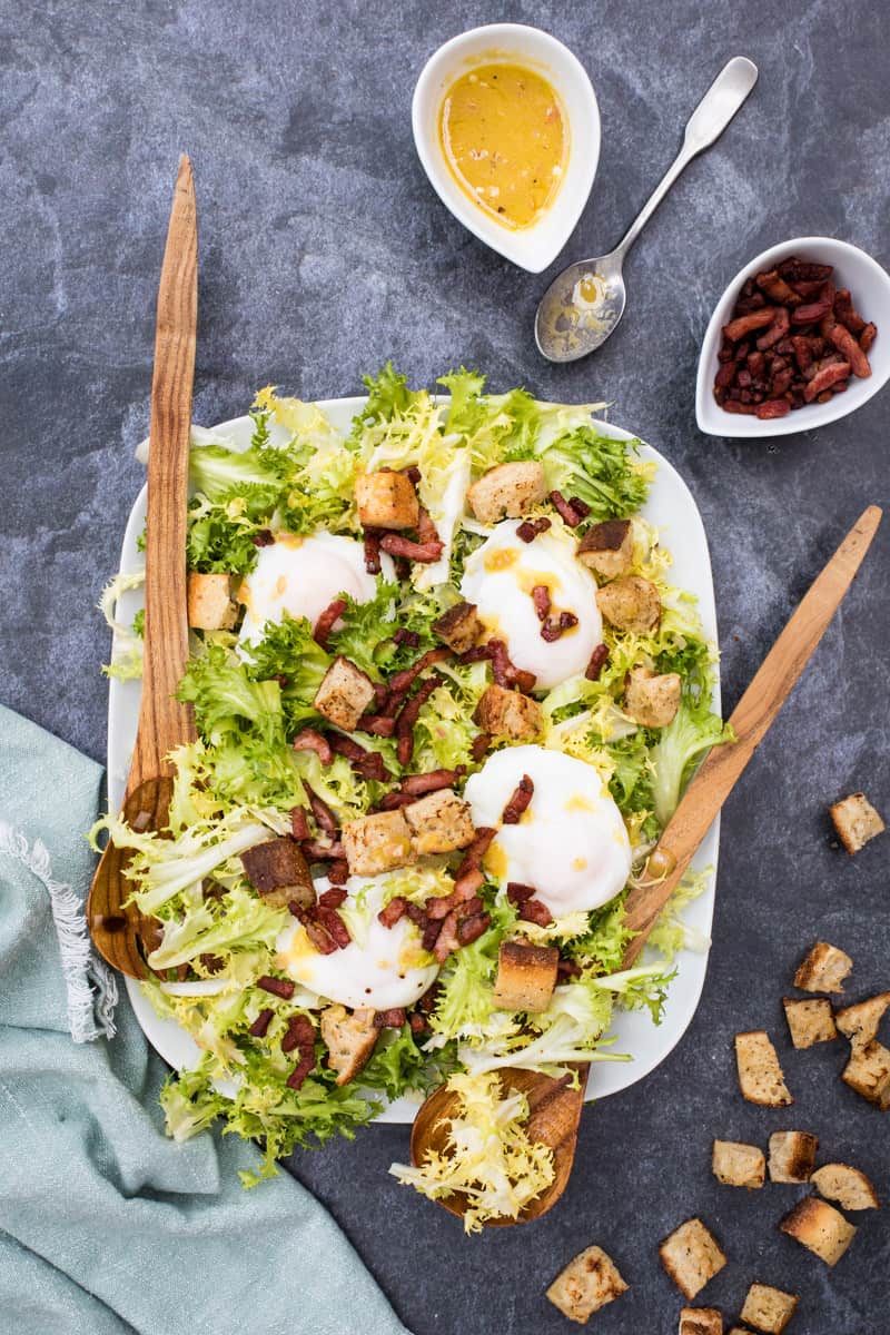 platter of frisee salad with bacon, poached eggs, and red wine vinaigrette with serving utensils and napkin