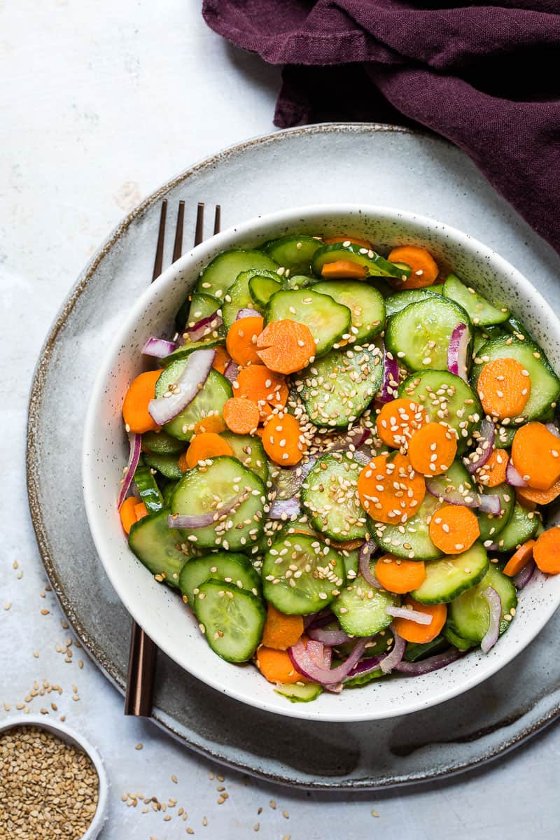 cucumber carrot salad in serving bowl with fork, napkin, and bowl of toasted sesame seeds